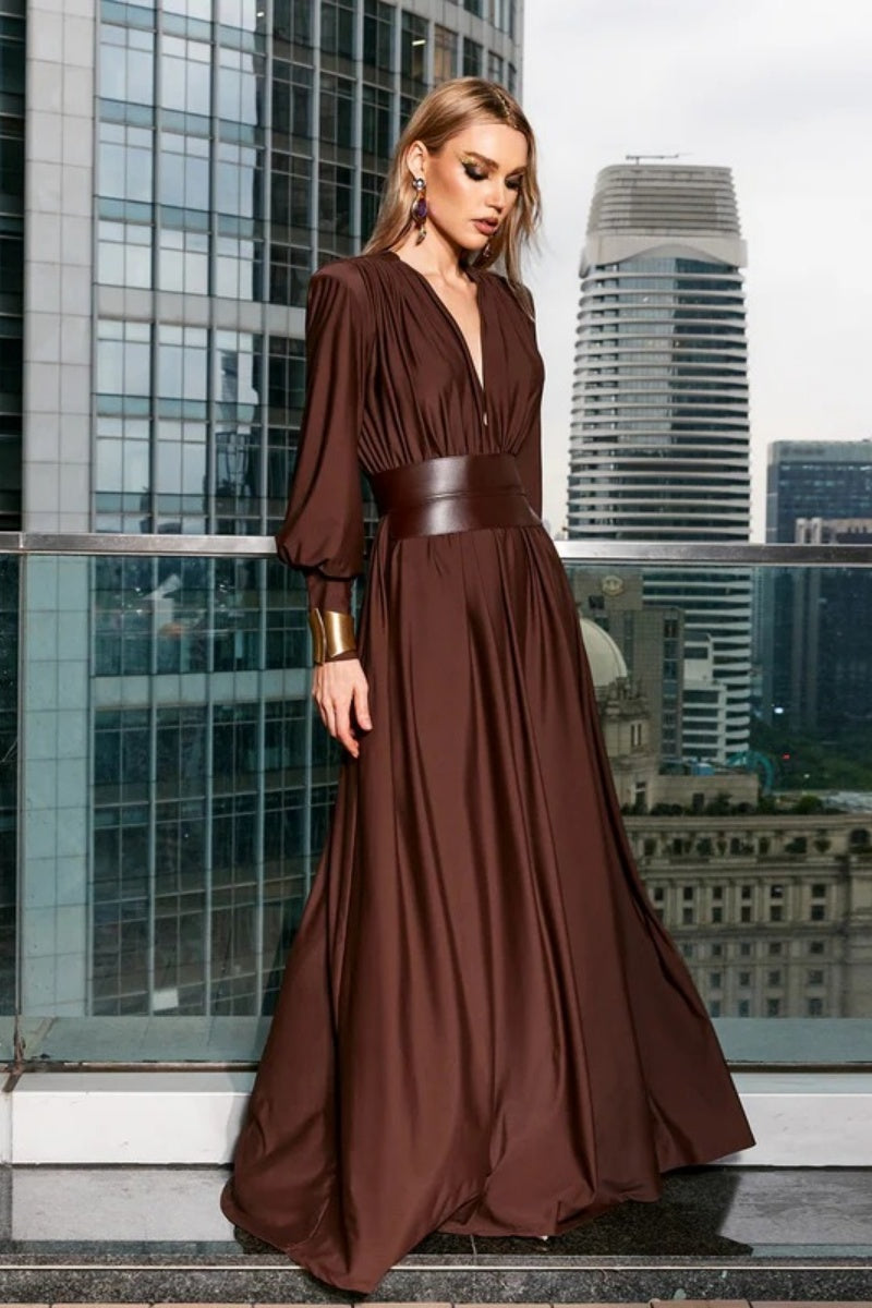 Brown - A woman wearing a chocolate brown satin maxi dress with a plunging V-neckline, ruched bodice, wide structured waist panel, and long bishop sleeves, standing against a city skyline background.