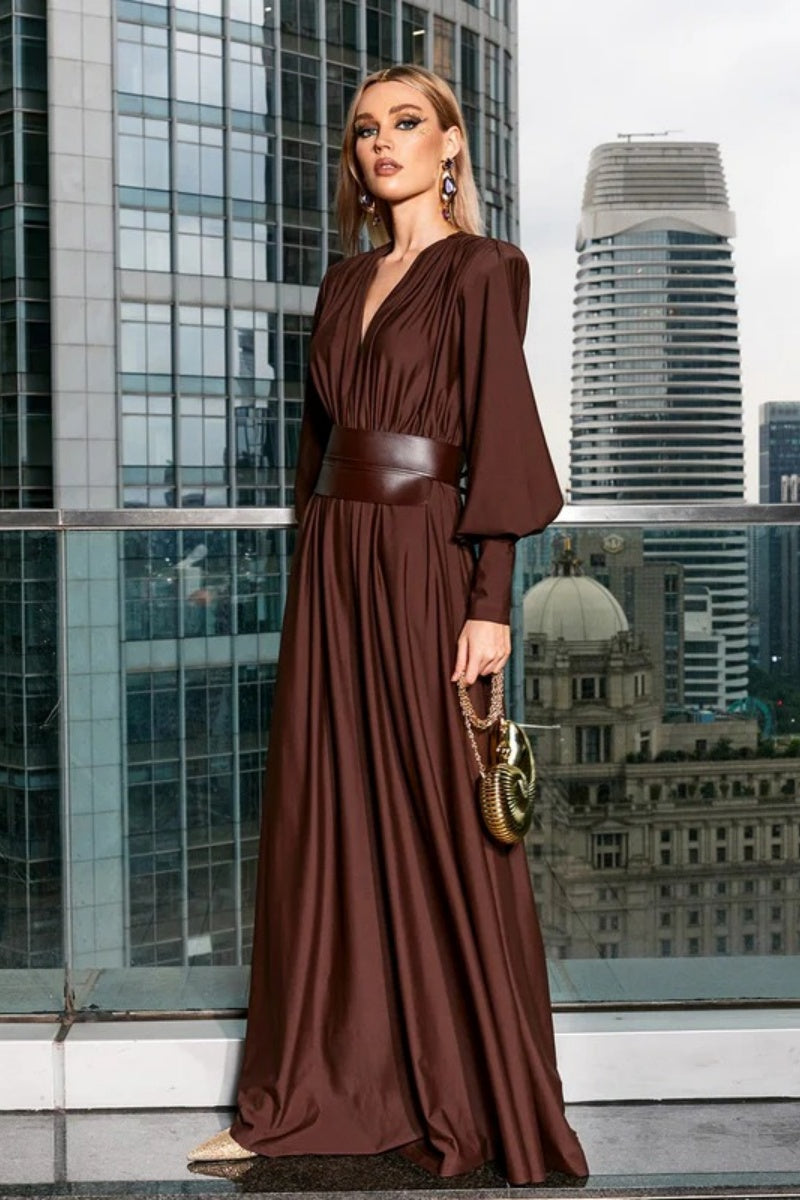Brown - A woman wearing a chocolate brown satin maxi dress with a plunging V-neckline, ruched bodice, wide structured waist panel, and long bishop sleeves, standing against a city skyline background.