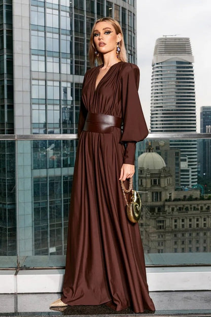 Brown - A woman wearing a chocolate brown satin maxi dress with a plunging V-neckline, ruched bodice, wide structured waist panel, and long bishop sleeves, standing against a city skyline background.