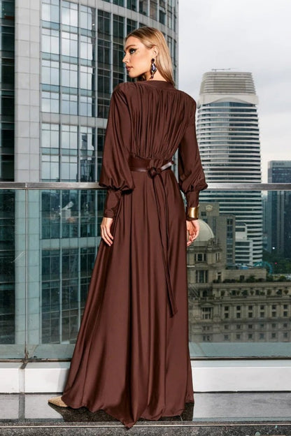 Brown - A woman wearing a chocolate brown satin maxi dress with a plunging V-neckline, ruched bodice, wide structured waist panel, and long bishop sleeves, standing against a city skyline background.