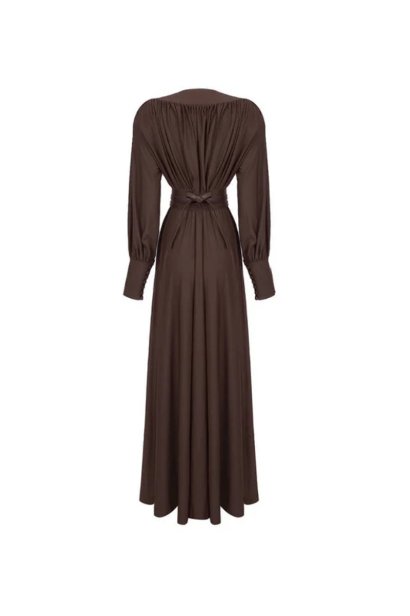 Brown - A woman wearing a chocolate brown satin maxi dress with a plunging V-neckline, ruched bodice, wide structured waist panel, and long bishop sleeves, standing against a city skyline background.