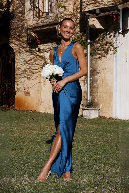 Blue - A woman wearing a bronze-coloured satin maxi dress with a plunging V-neckline, twist-front waist detail, and a front split, standing on a rustic stone balcony.