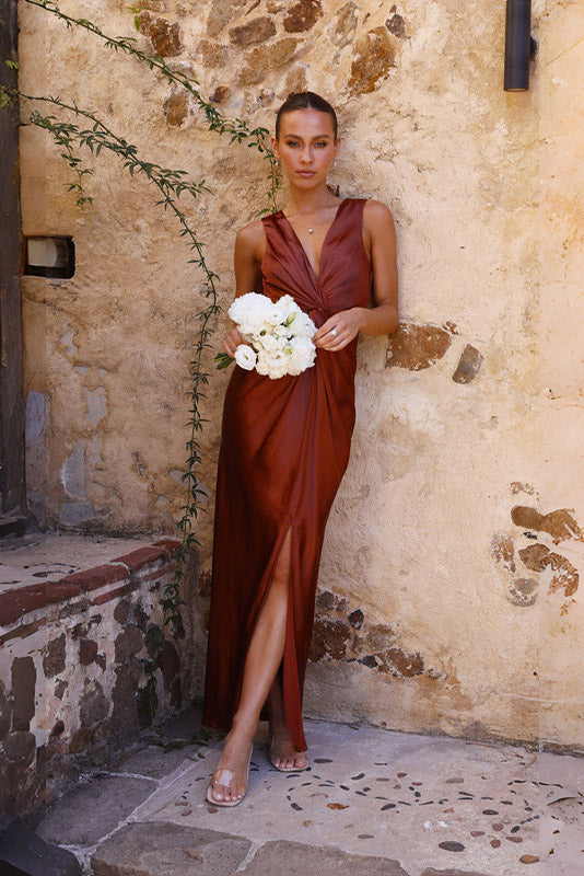 Coffee - A woman wearing a bronze-coloured satin maxi dress with a plunging V-neckline, twist-front waist detail, and a front split, standing on a rustic stone balcony.