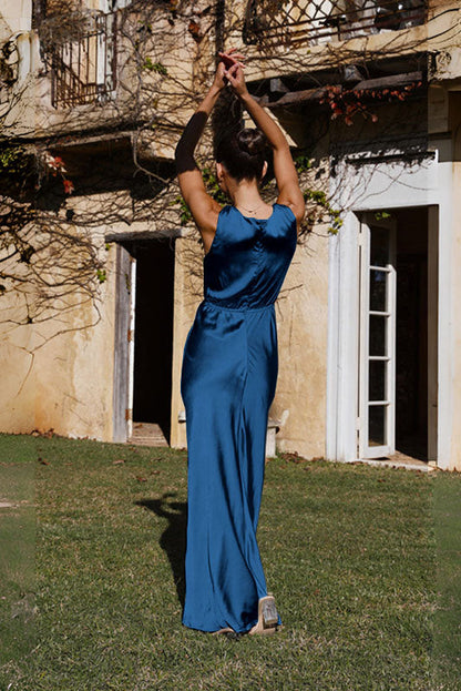 Coffee - A woman wearing a bronze-coloured satin maxi dress with a plunging V-neckline, twist-front waist detail, and a front split, standing on a rustic stone balcony.
