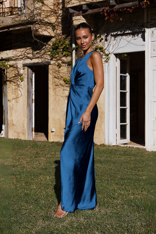 Coffee - A woman wearing a bronze-coloured satin maxi dress with a plunging V-neckline, twist-front waist detail, and a front split, standing on a rustic stone balcony.