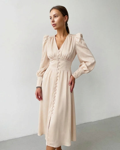 Beige - A woman wearing a taupe-coloured satin midi dress featuring a V-neckline, a row of self-covered buttons down the front, a cinched waistband, and long bishop sleeves with extended buttoned cuffs.