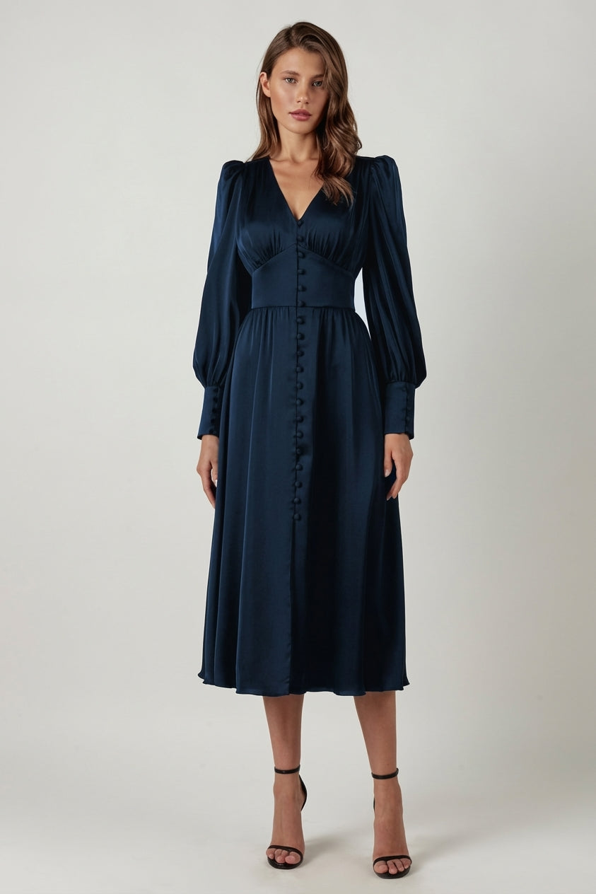 Blue - A woman wearing a taupe-coloured satin midi dress featuring a V-neckline, a row of self-covered buttons down the front, a cinched waistband, and long bishop sleeves with extended buttoned cuffs.