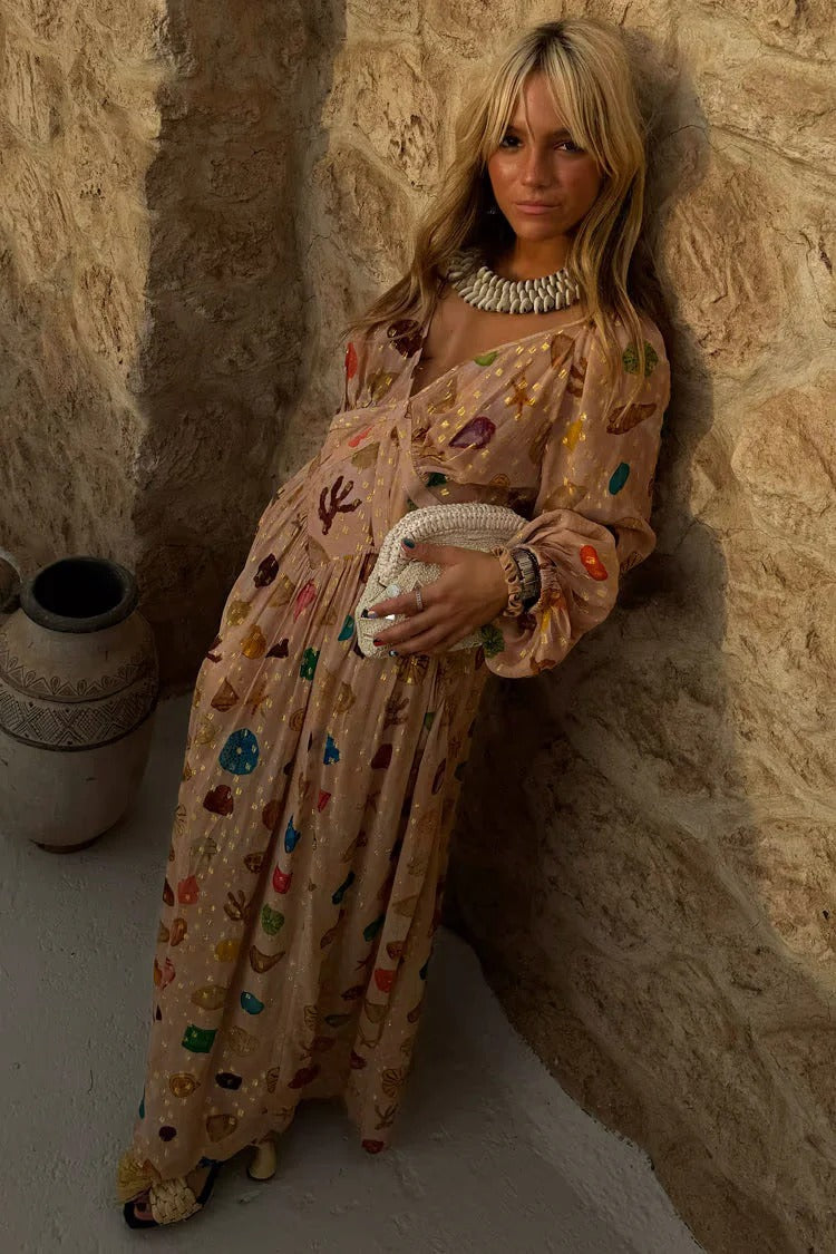 A woman leans against a textured stone wall wearing a peach-coloured maxi dress featuring a multi-coloured seashell motif, gold Lurex flecks, a plunging V-neckline, and long billowy sleeves.