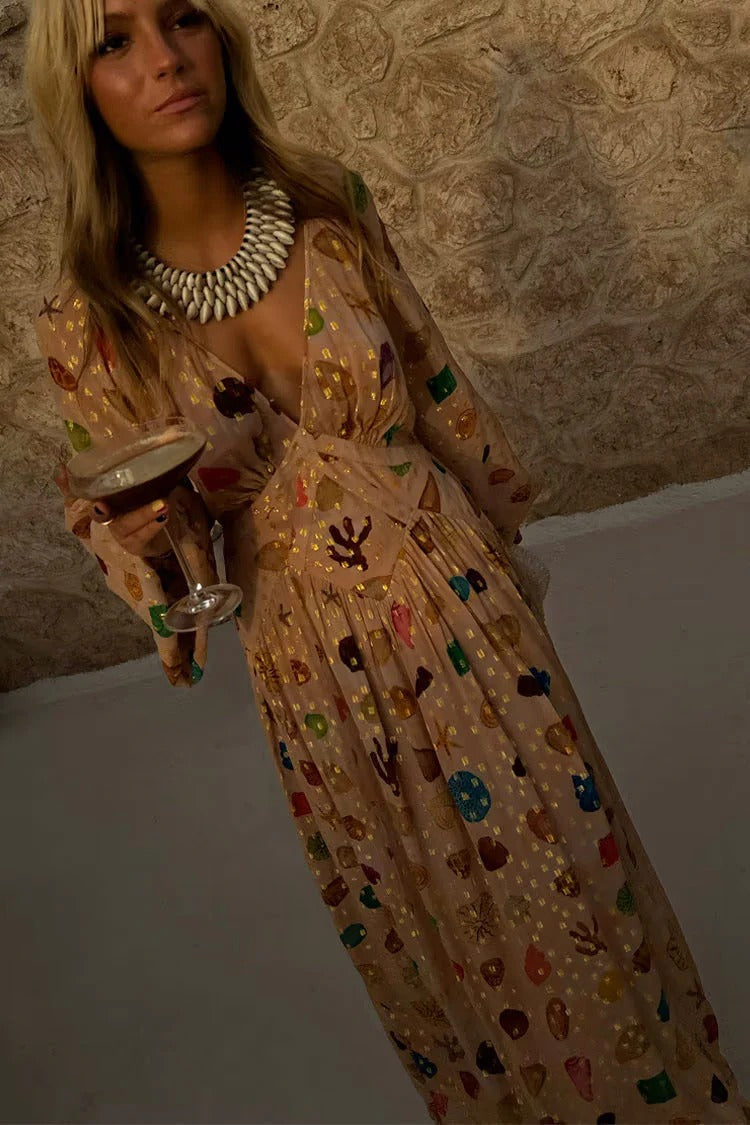 A woman leans against a textured stone wall wearing a peach-coloured maxi dress featuring a multi-coloured seashell motif, gold Lurex flecks, a plunging V-neckline, and long billowy sleeves.