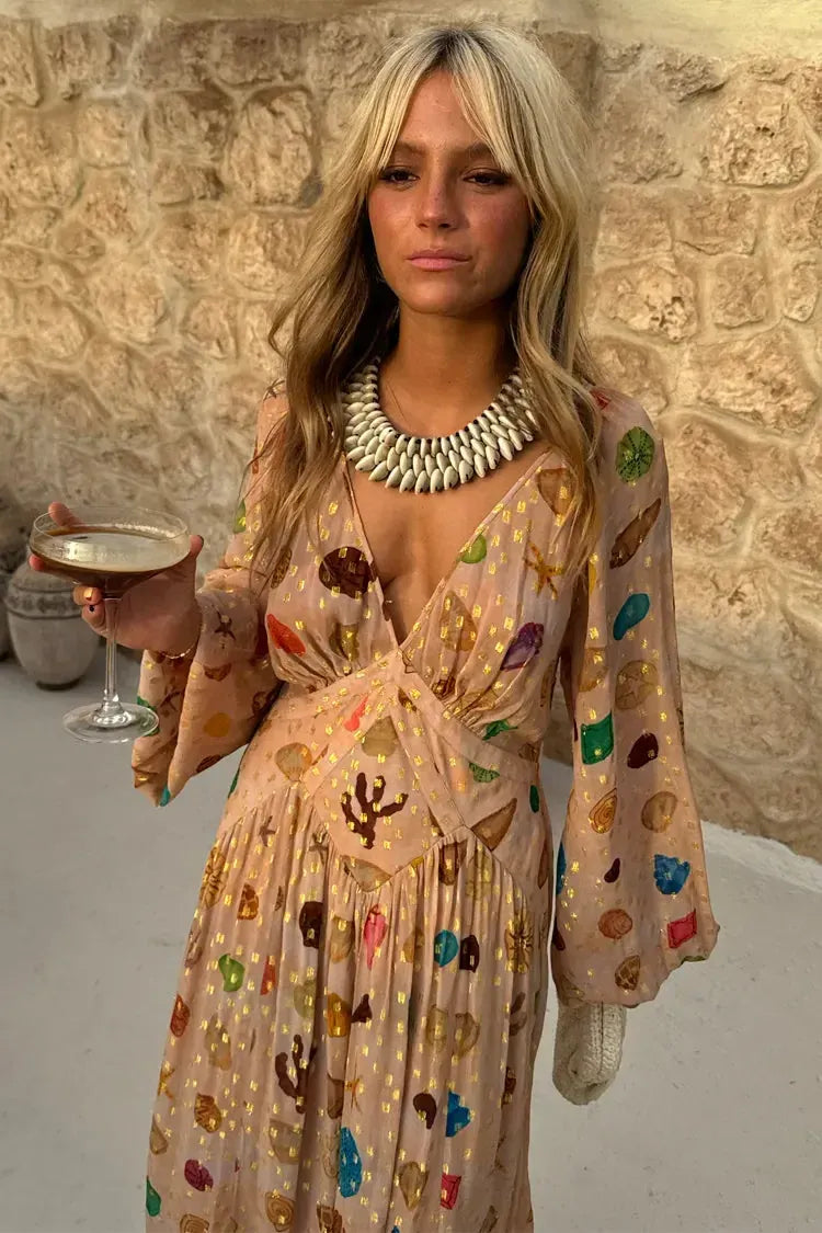 A woman leans against a textured stone wall wearing a peach-coloured maxi dress featuring a multi-coloured seashell motif, gold Lurex flecks, a plunging V-neckline, and long billowy sleeves.