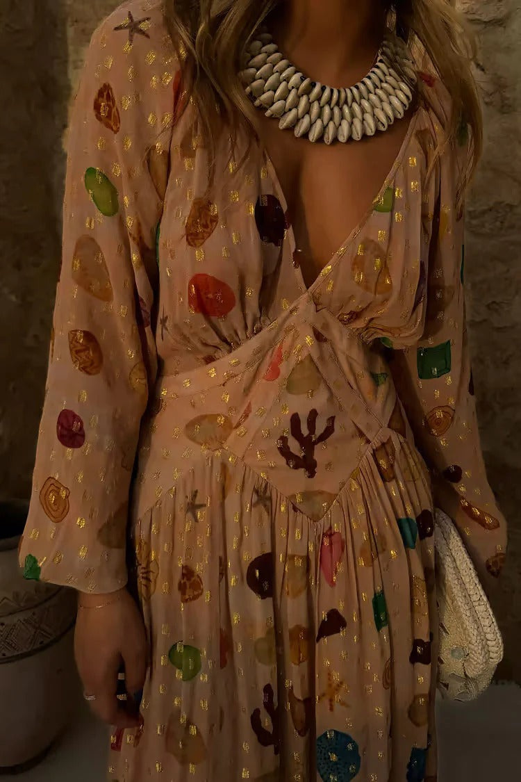 A woman leans against a textured stone wall wearing a peach-coloured maxi dress featuring a multi-coloured seashell motif, gold Lurex flecks, a plunging V-neckline, and long billowy sleeves.