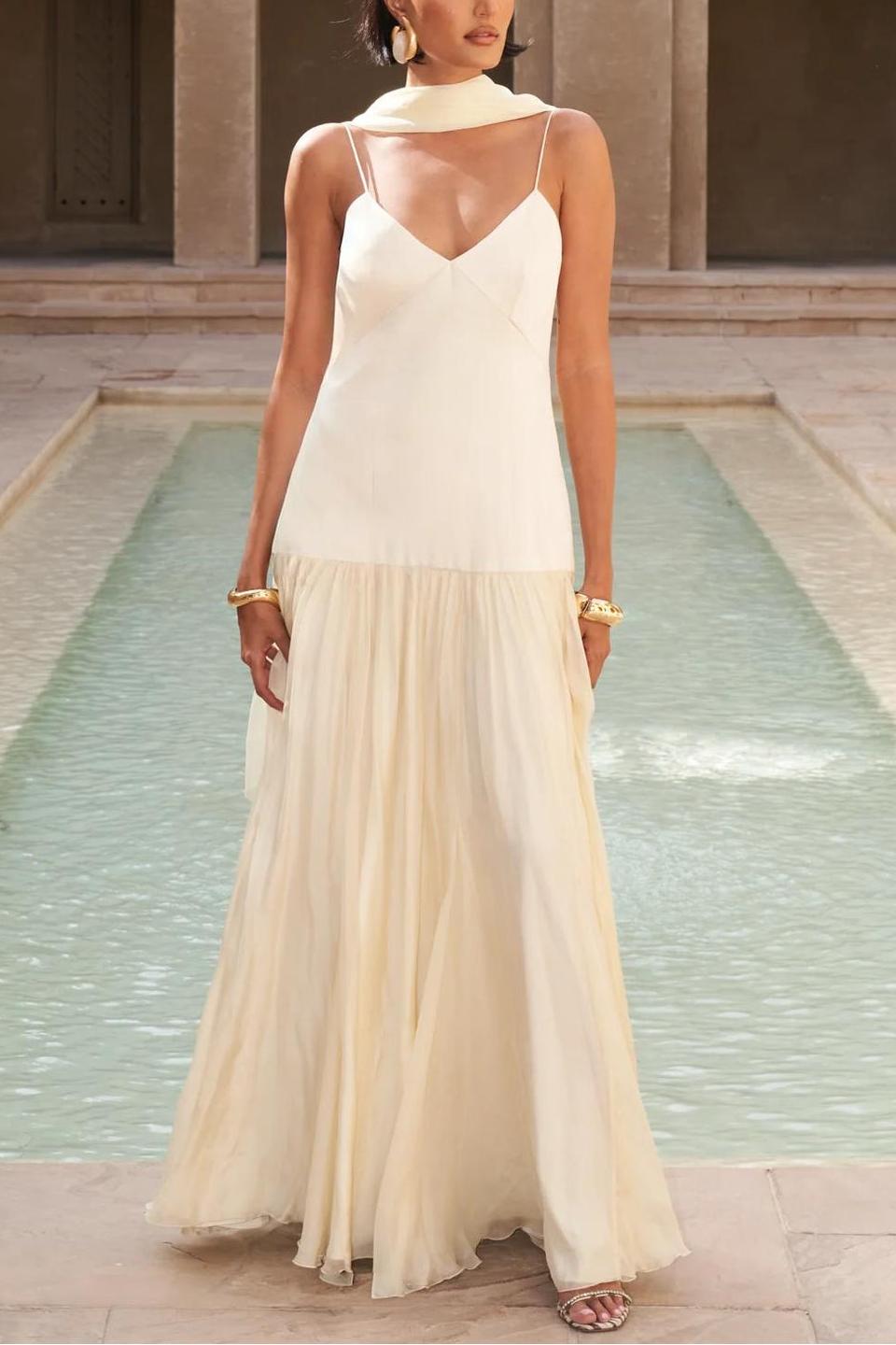 Women's elegant drop-waist maxi dress featuring a smooth cream V-neck bodice, delicate spaghetti straps, and a voluminous pale yellow sheer flowing skirt, styled with a matching neck scarf in a sunlit courtyard.