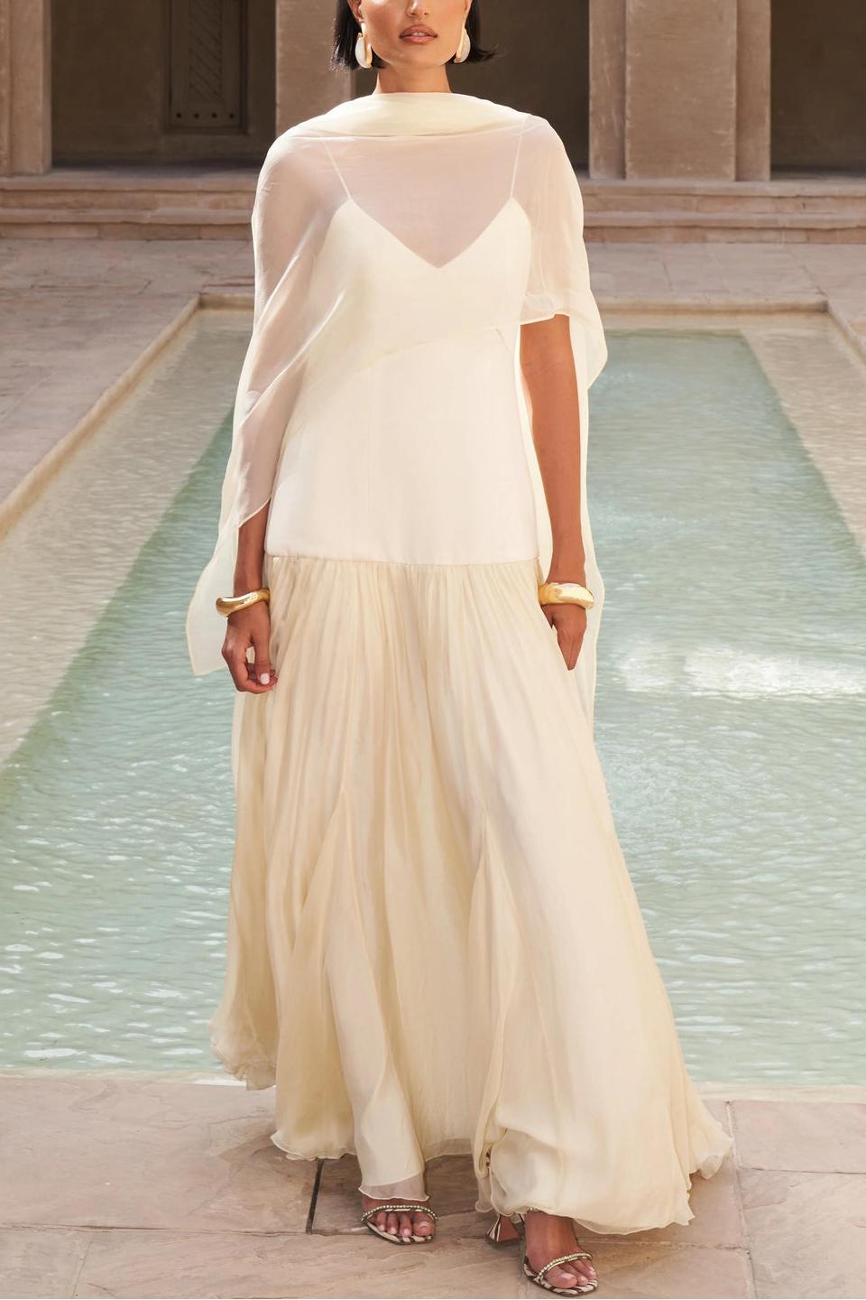 Women's elegant drop-waist maxi dress featuring a smooth cream V-neck bodice, delicate spaghetti straps, and a voluminous pale yellow sheer flowing skirt, styled with a matching neck scarf in a sunlit courtyard.