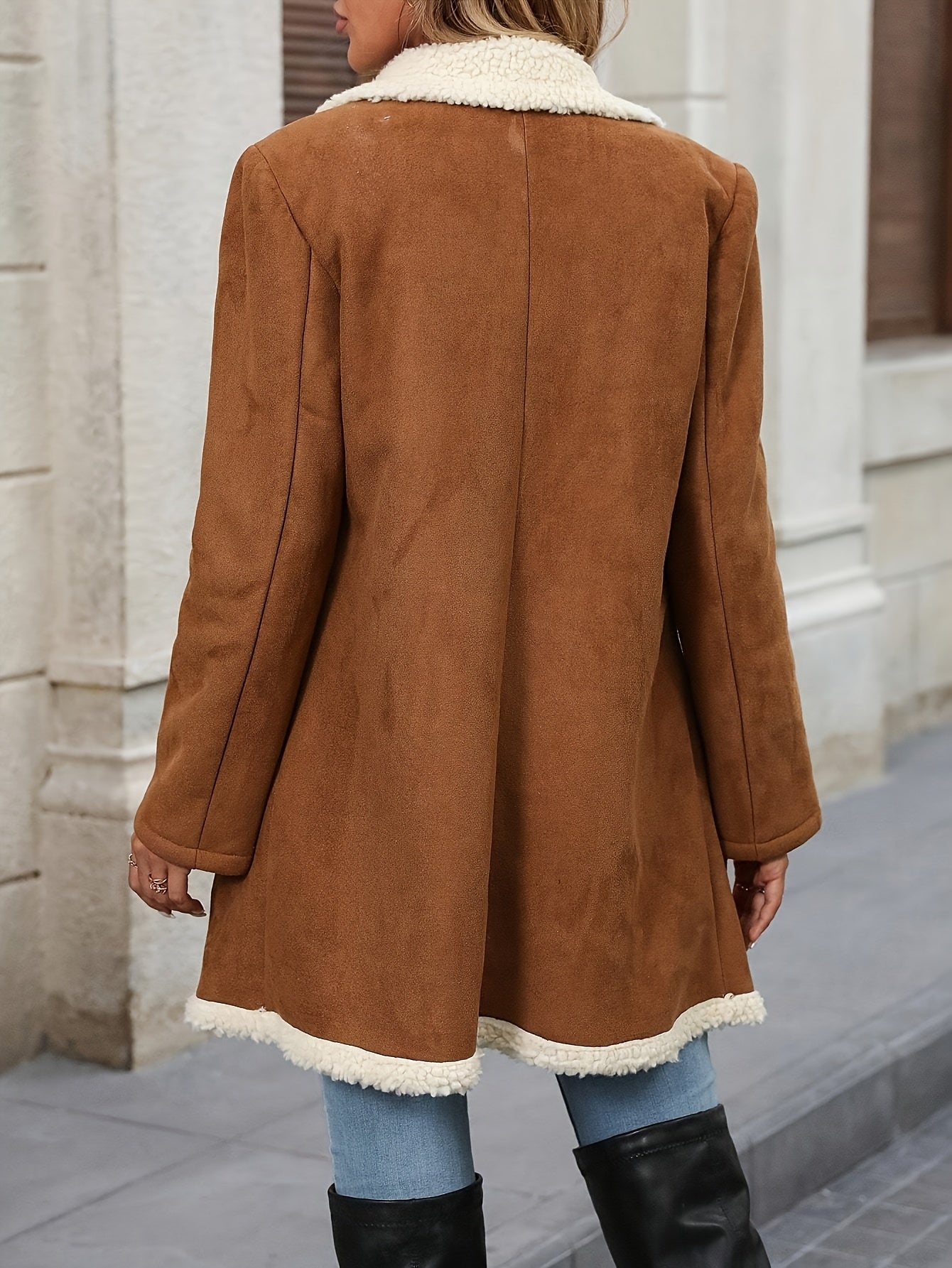 A woman wearing a mid-length tan suede-look coat with a plush cream sherpa lining on the collar, lapels, and hem, featuring a double-breasted button closure.