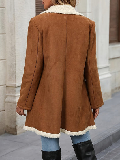 A woman wearing a mid-length tan suede-look coat with a plush cream sherpa lining on the collar, lapels, and hem, featuring a double-breasted button closure.