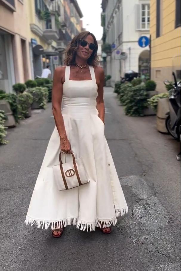 A woman stands on a cobblestone street wearing a white, square-neck maxi dress with a fitted bodice, full skirt, and a fringed hem.