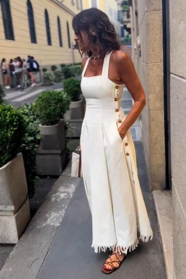 White - A woman stands on a cobblestone street wearing a white, square-neck maxi dress with a fitted bodice, full skirt, and a fringed hem.