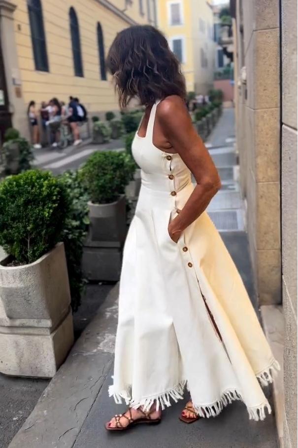 White - A woman stands on a cobblestone street wearing a white, square-neck maxi dress with a fitted bodice, full skirt, and a fringed hem.