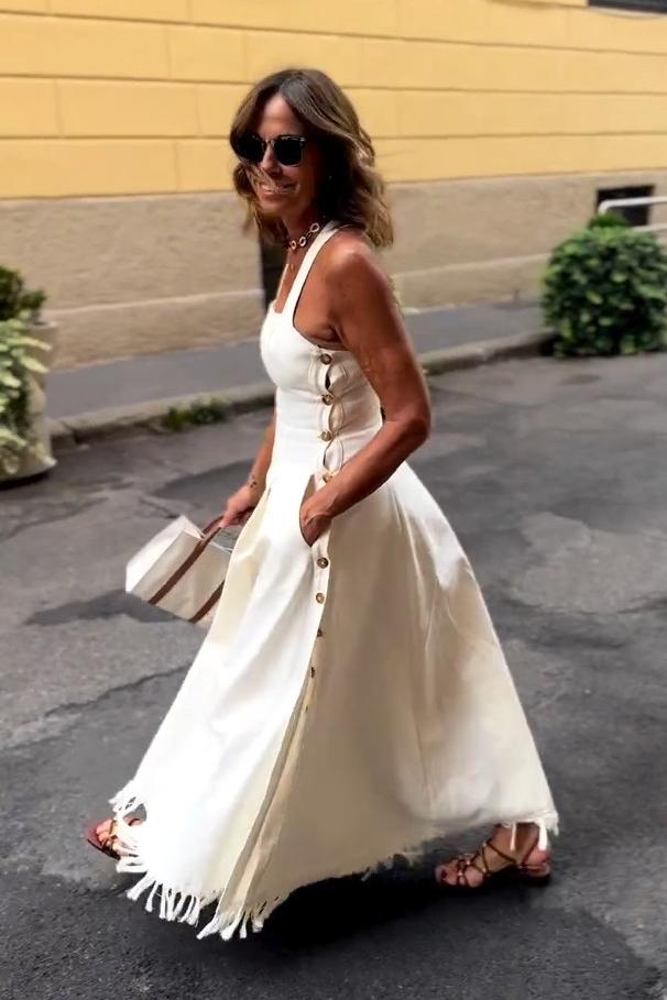 White - A woman stands on a cobblestone street wearing a white, square-neck maxi dress with a fitted bodice, full skirt, and a fringed hem.