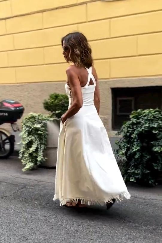 White - A woman stands on a cobblestone street wearing a white, square-neck maxi dress with a fitted bodice, full skirt, and a fringed hem.