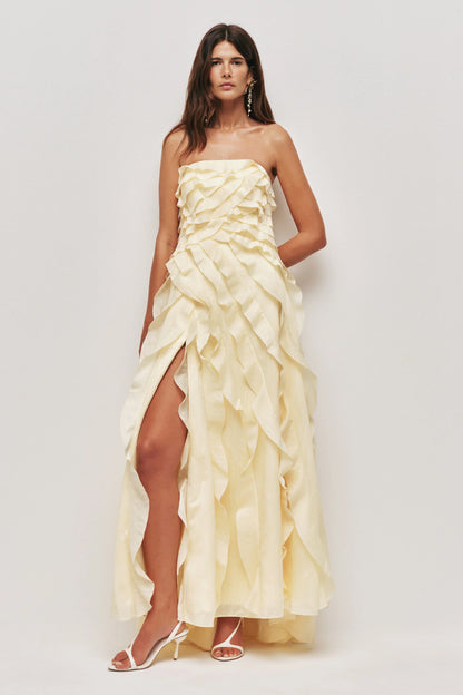 A woman wearing a pale yellow, strapless maxi dress featuring all-over cascading ruffles and a high side slit, styled with white strappy heels.