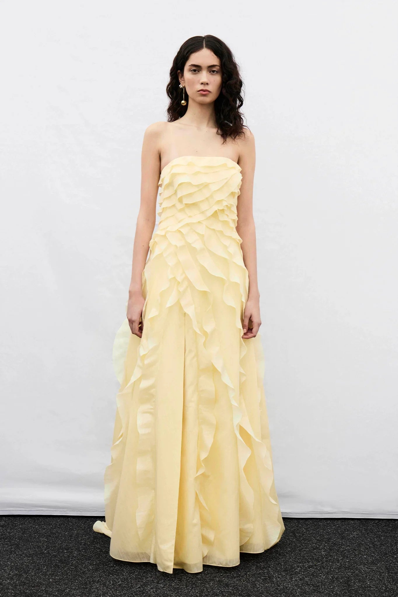 A woman wearing a pale yellow, strapless maxi dress featuring all-over cascading ruffles and a high side slit, styled with white strappy heels.