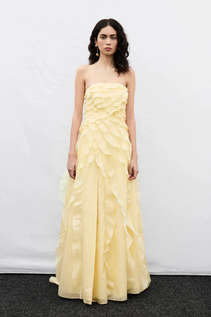 A woman wearing a pale yellow, strapless maxi dress featuring all-over cascading ruffles and a high side slit, styled with white strappy heels.