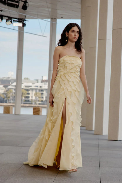 A woman wearing a pale yellow, strapless maxi dress featuring all-over cascading ruffles and a high side slit, styled with white strappy heels.