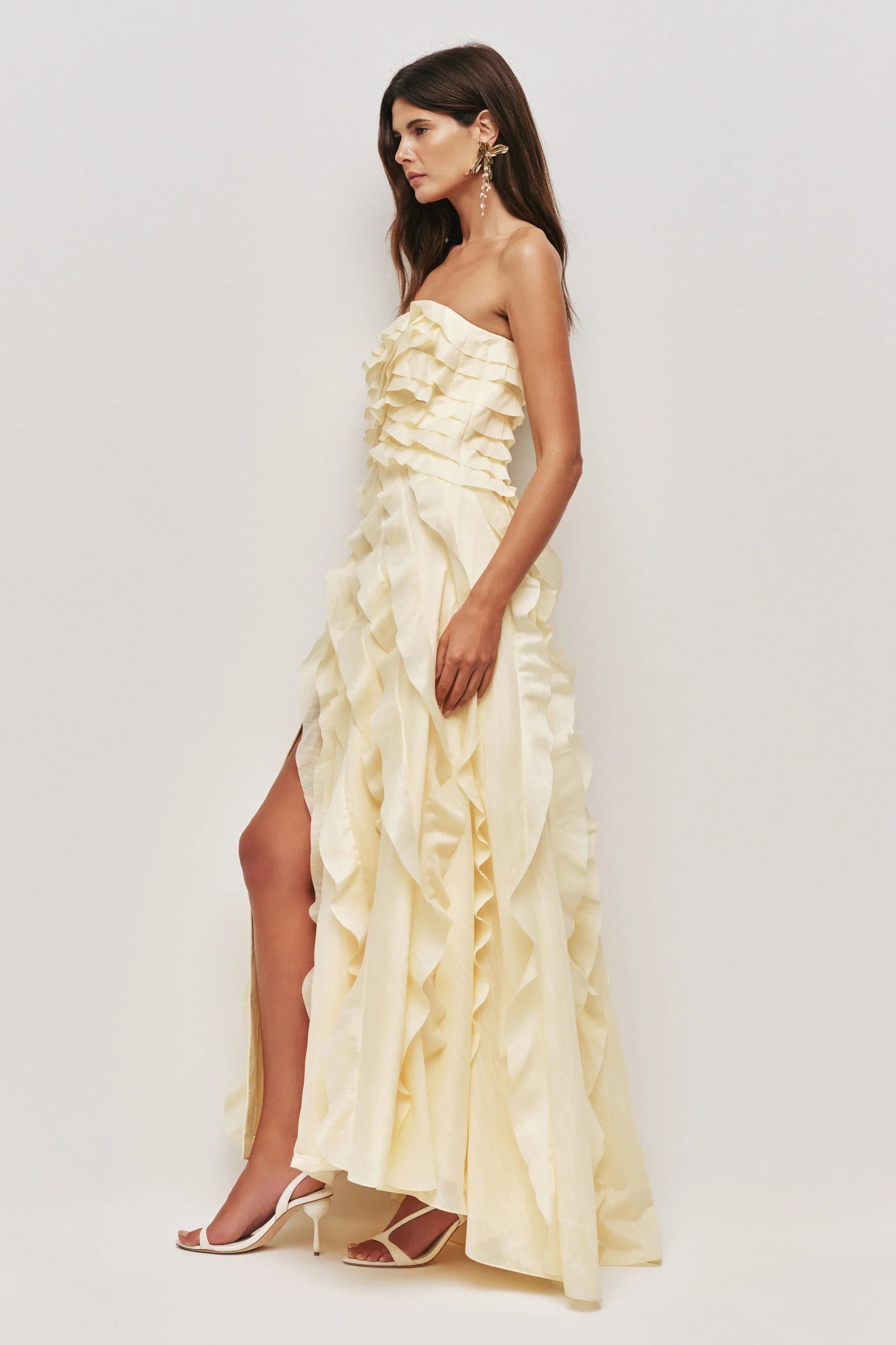 A woman wearing a pale yellow, strapless maxi dress featuring all-over cascading ruffles and a high side slit, styled with white strappy heels.