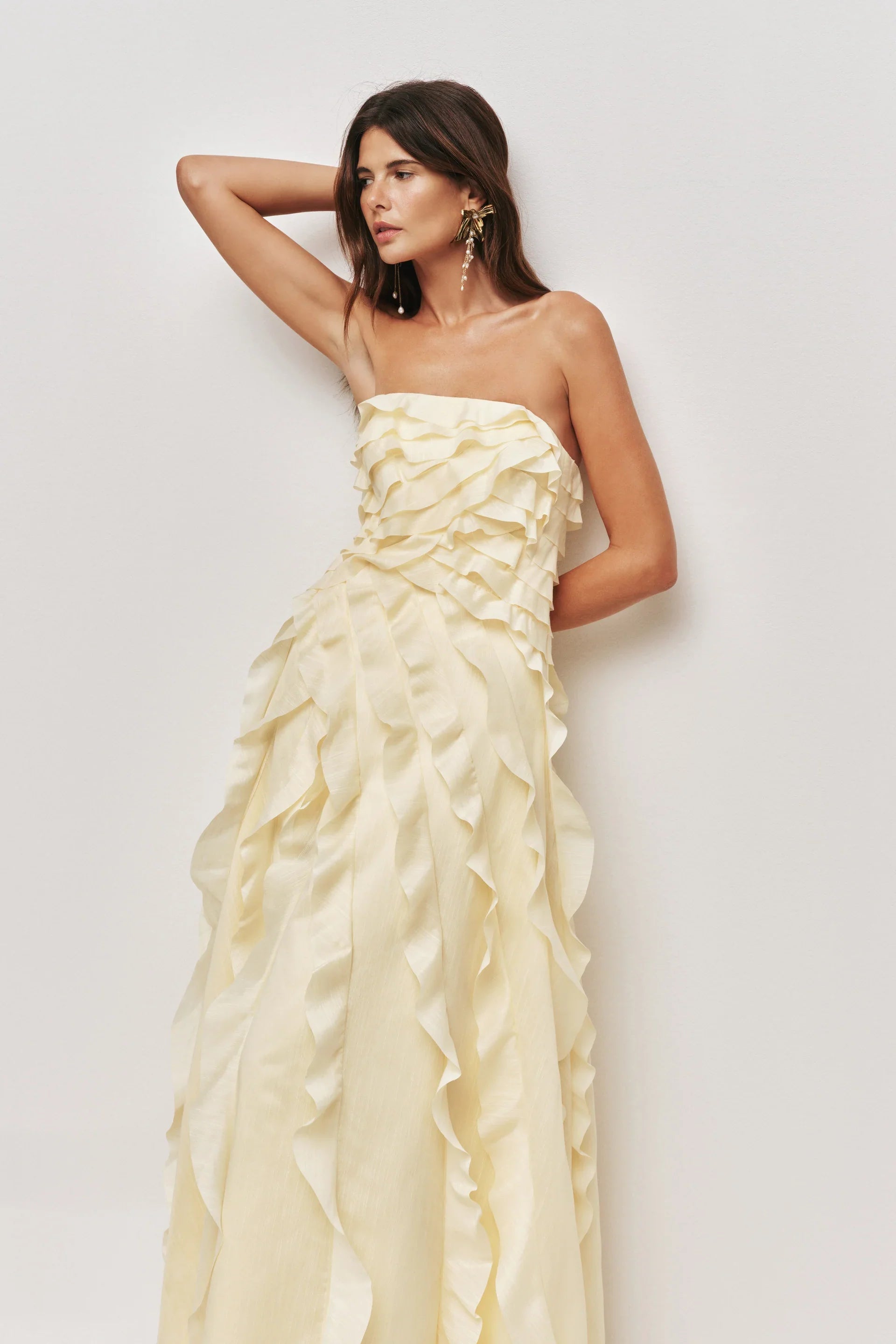 A woman wearing a pale yellow, strapless maxi dress featuring all-over cascading ruffles and a high side slit, styled with white strappy heels.