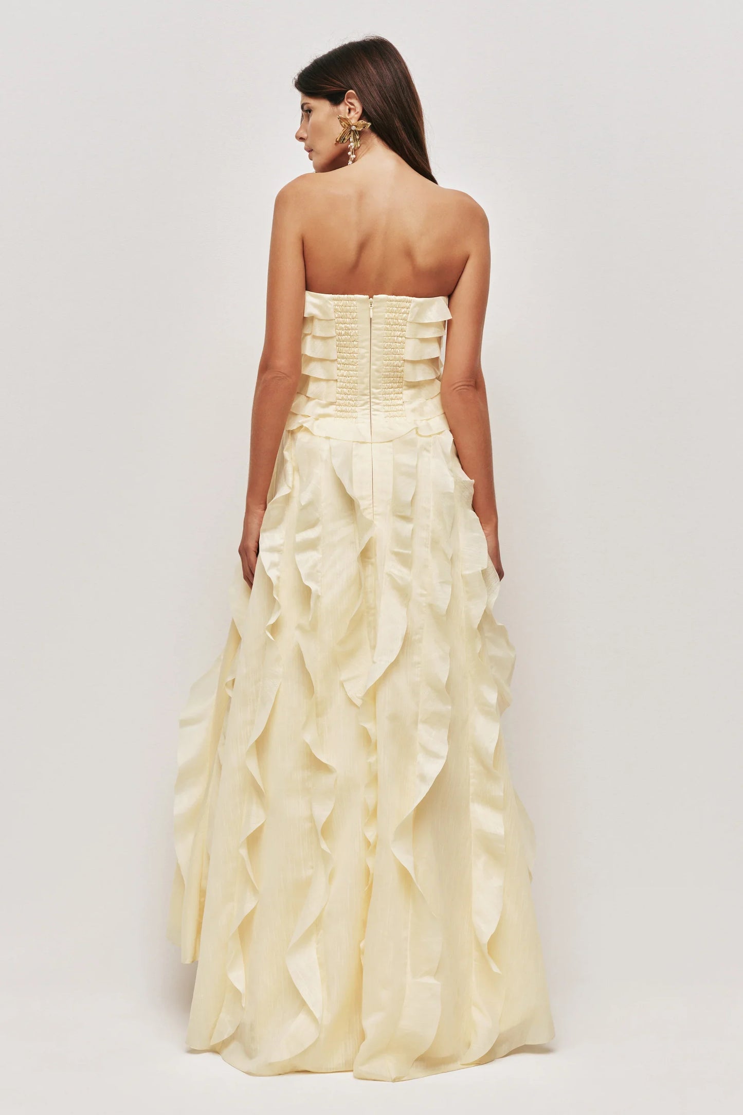 A woman wearing a pale yellow, strapless maxi dress featuring all-over cascading ruffles and a high side slit, styled with white strappy heels.