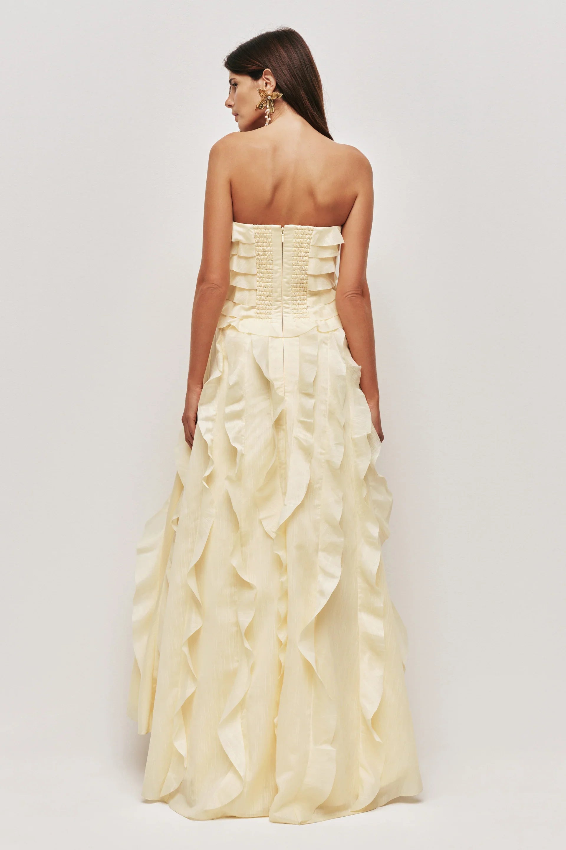 A woman wearing a pale yellow, strapless maxi dress featuring all-over cascading ruffles and a high side slit, styled with white strappy heels.