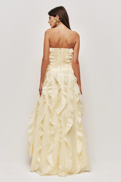 A woman wearing a pale yellow, strapless maxi dress featuring all-over cascading ruffles and a high side slit, styled with white strappy heels.