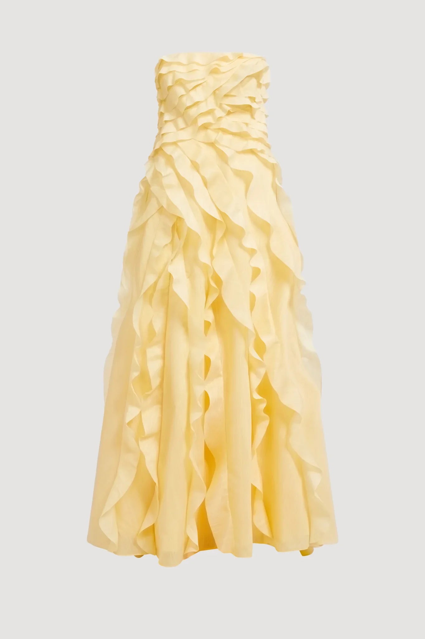 A woman wearing a pale yellow, strapless maxi dress featuring all-over cascading ruffles and a high side slit, styled with white strappy heels.