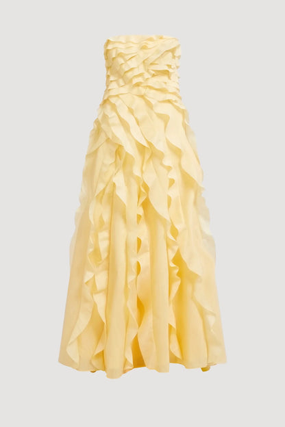 A woman wearing a pale yellow, strapless maxi dress featuring all-over cascading ruffles and a high side slit, styled with white strappy heels.