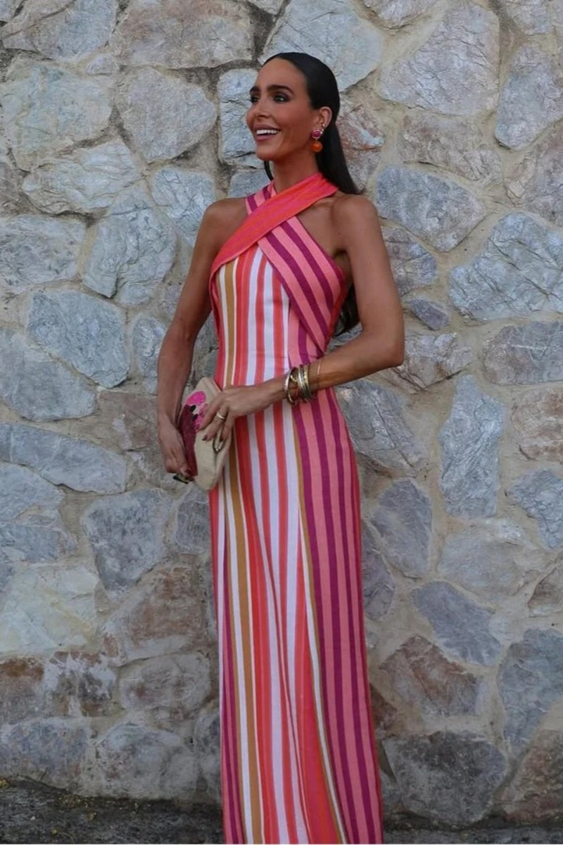 Red - A woman wearing a vibrant pink, orange, and white vertically striped maxi dress with a crossover halter neckline, posing against a rustic stone wall.