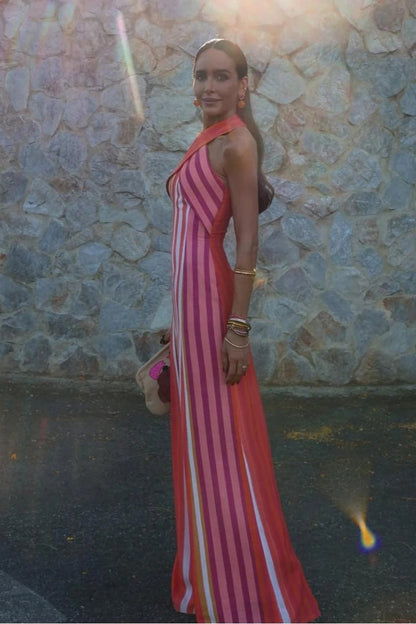 Red - A woman wearing a vibrant pink, orange, and white vertically striped maxi dress with a crossover halter neckline, posing against a rustic stone wall.