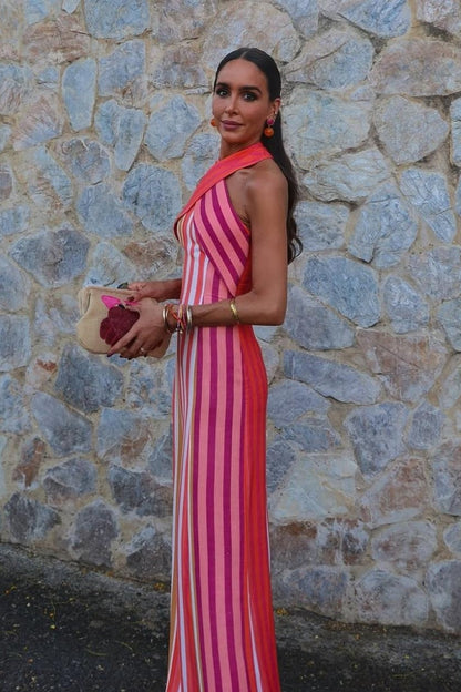 Red - A woman wearing a vibrant pink, orange, and white vertically striped maxi dress with a crossover halter neckline, posing against a rustic stone wall.