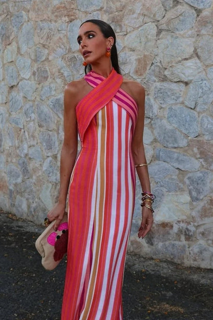 Red - A woman wearing a vibrant pink, orange, and white vertically striped maxi dress with a crossover halter neckline, posing against a rustic stone wall.
