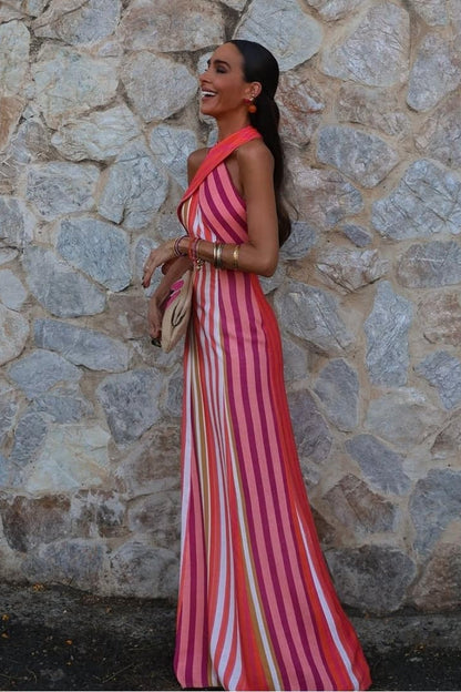 Red - A woman wearing a vibrant pink, orange, and white vertically striped maxi dress with a crossover halter neckline, posing against a rustic stone wall.