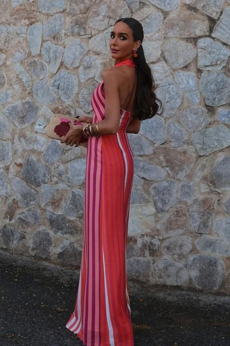 Red - A woman wearing a vibrant pink, orange, and white vertically striped maxi dress with a crossover halter neckline, posing against a rustic stone wall.
