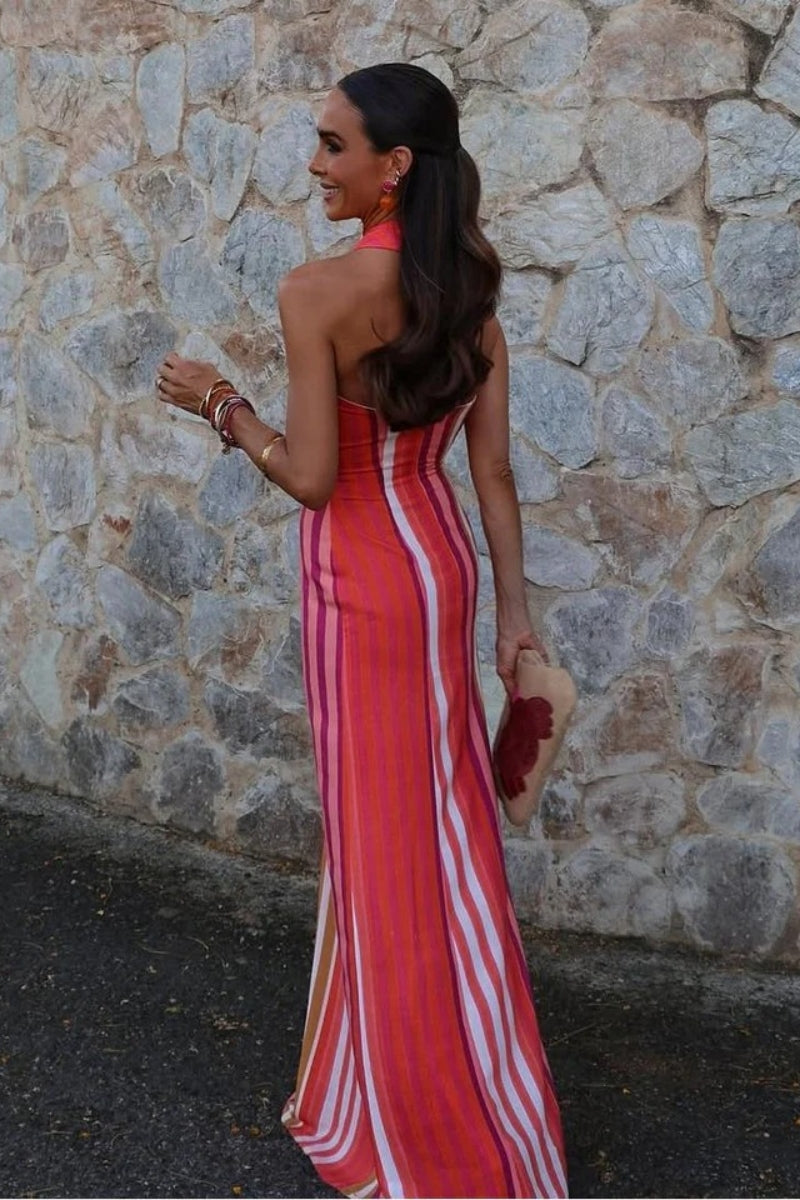 Red - A woman wearing a vibrant pink, orange, and white vertically striped maxi dress with a crossover halter neckline, posing against a rustic stone wall.
