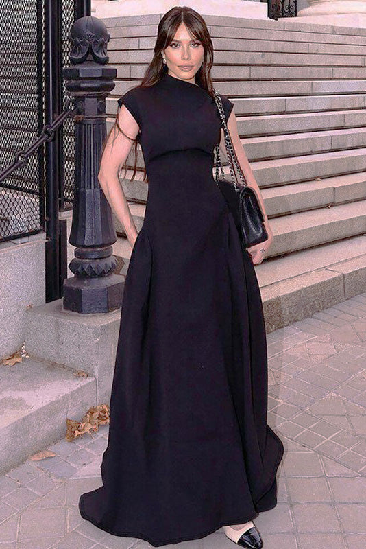 A woman models a formal white sleeveless maxi dress featuring a high neckline, fitted bodice, and dramatic voluminous skirt with structured hip gathering, standing on brick steps.
