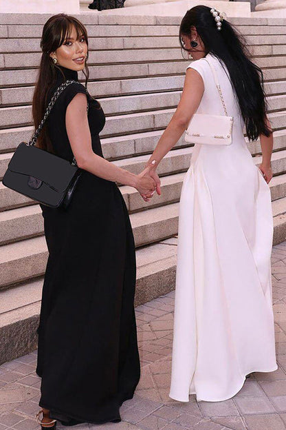 A woman models a formal white sleeveless maxi dress featuring a high neckline, fitted bodice, and dramatic voluminous skirt with structured hip gathering, standing on brick steps.
