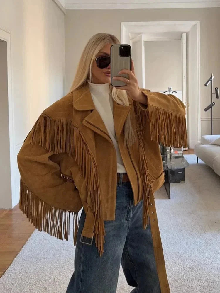 A woman wearing an oversized tan suede jacket featuring extensive fringe detailing along the chest and sleeves, styled with a white turtleneck and blue jeans.