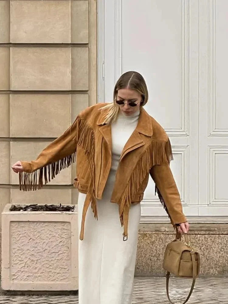A woman wearing an oversized tan suede jacket featuring extensive fringe detailing along the chest and sleeves, styled with a white turtleneck and blue jeans.