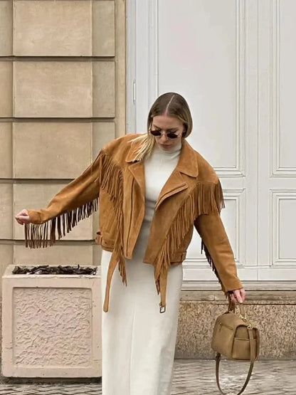 A woman wearing an oversized tan suede jacket featuring extensive fringe detailing along the chest and sleeves, styled with a white turtleneck and blue jeans.