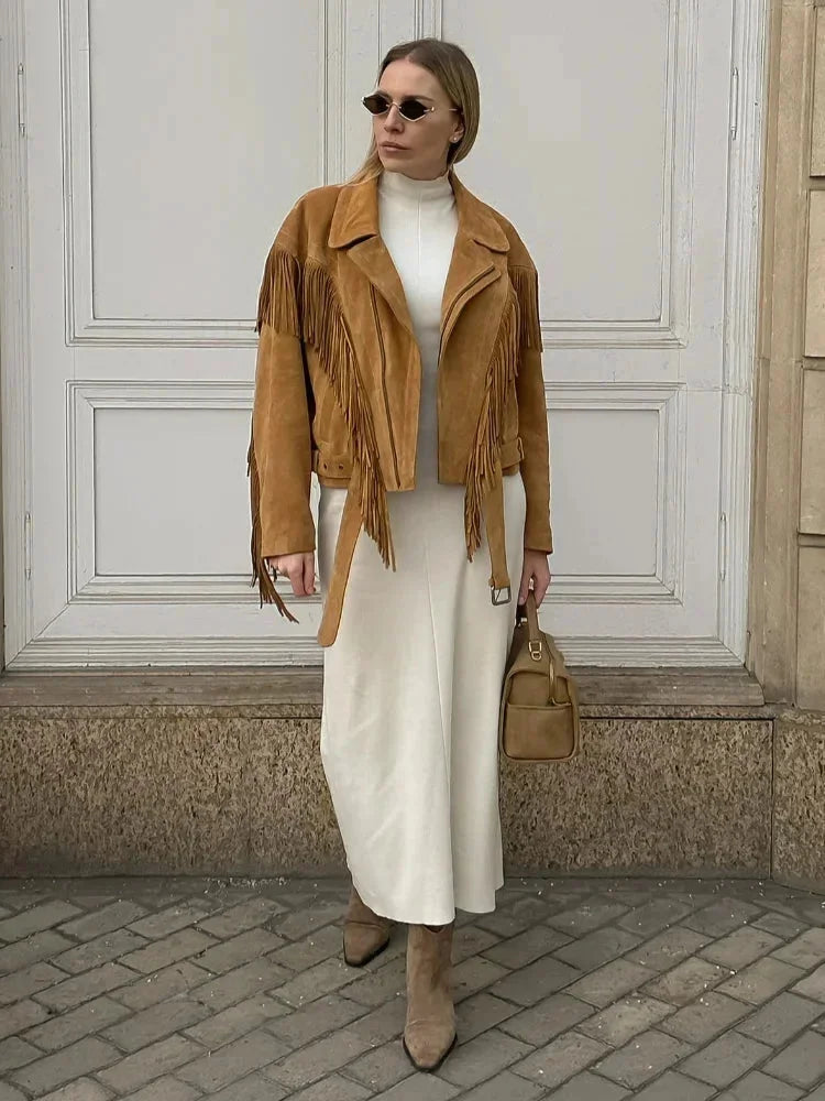 A woman wearing an oversized tan suede jacket featuring extensive fringe detailing along the chest and sleeves, styled with a white turtleneck and blue jeans.