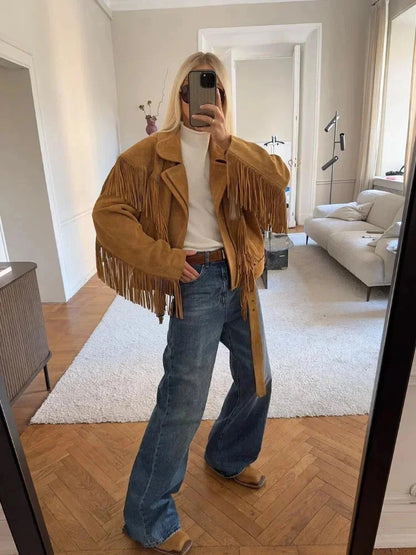 A woman wearing an oversized tan suede jacket featuring extensive fringe detailing along the chest and sleeves, styled with a white turtleneck and blue jeans.