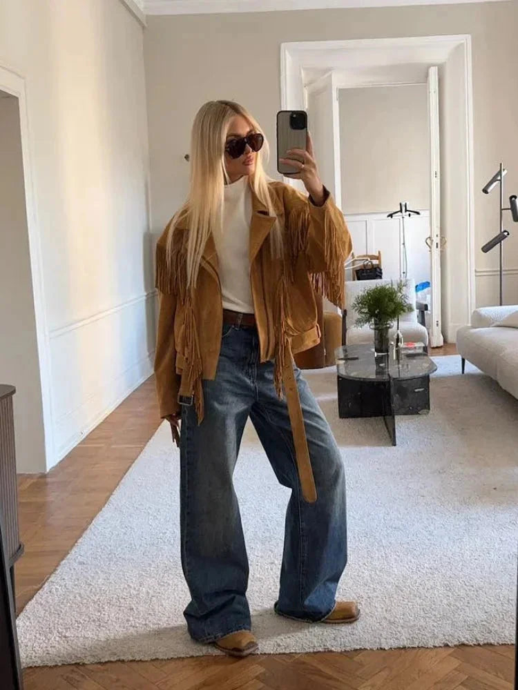 A woman wearing an oversized tan suede jacket featuring extensive fringe detailing along the chest and sleeves, styled with a white turtleneck and blue jeans.